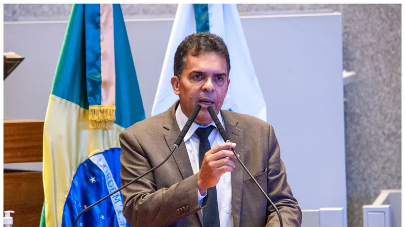 A fotografia mostra o deputado Ricardo Vale discursando em uma tribuna. Ele é um homem de meia-idade, com cabelos curtos e grisalhos, vestindo terno cinza, camisa branca e gravata escura. Posicionado atrás de um púlpito de madeira com dois microfones à sua frente, ele gesticula com a mão direita parcialmente erguida na altura do peito. Ao fundo, estão posicionadas duas bandeiras: a do Brasil à esquerda, com o lema "Ordem e Progresso" parcialmente visível, e uma bandeira branca à direita. Sobre a extremidade esquerda do púlpito, há um pequeno frasco de álcool em gel.