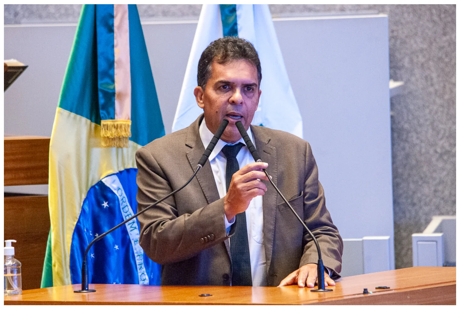 A fotografia mostra o deputado Ricardo Vale discursando em uma tribuna. Ele é um homem de meia-idade, com cabelos curtos e grisalhos, vestindo terno cinza, camisa branca e gravata escura. Posicionado atrás de um púlpito de madeira com dois microfones à sua frente, ele gesticula com a mão direita parcialmente erguida na altura do peito. Ao fundo, estão posicionadas duas bandeiras: a do Brasil à esquerda, com o lema "Ordem e Progresso" parcialmente visível, e uma bandeira branca à direita. Sobre a extremidade esquerda do púlpito, há um pequeno frasco de álcool em gel.