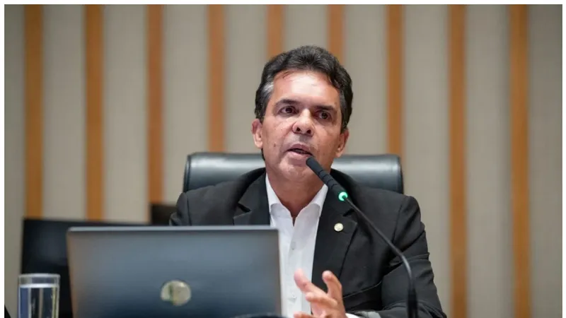 A imagem mostra o Deputado Ricardo Vale, sentado atrás de uma bancada de madeira. Ele está falando em um microfone e gesticula com a mão direita. À sua frente há um notebook prata aberto e um copo de água. O fundo é composto por painéis verticais xde madeira clara, sugerindo um ambiente de plenário ou sala de reuniões.