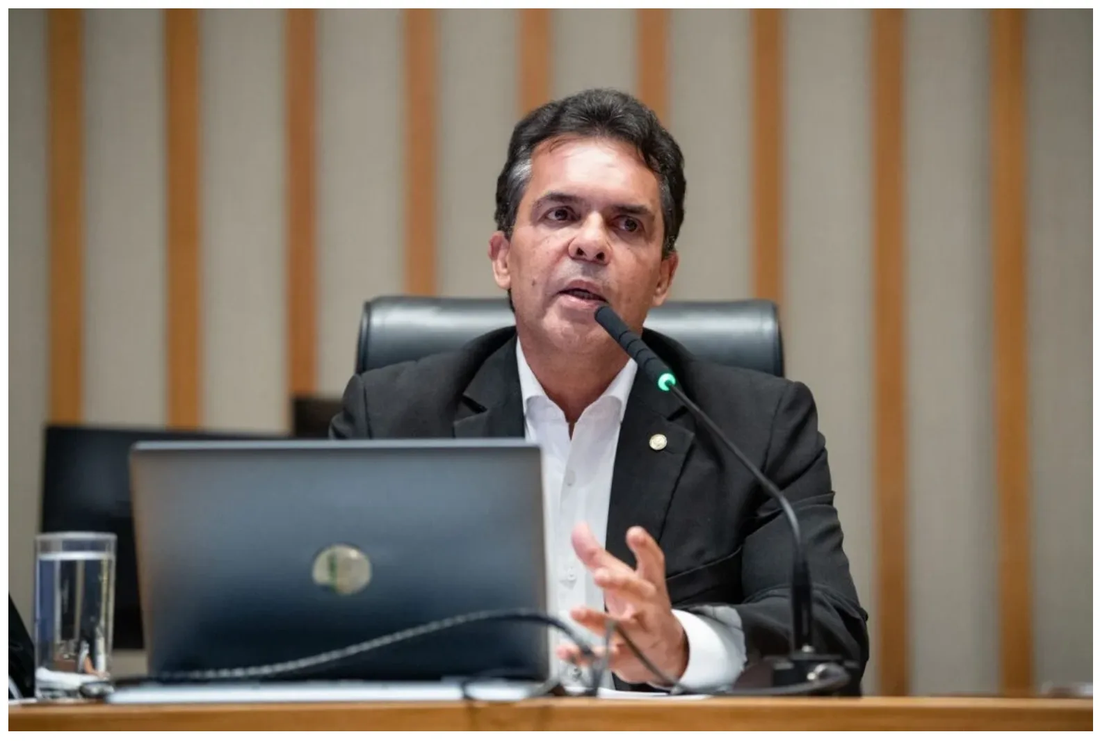 A imagem mostra o Deputado Ricardo Vale, sentado atrás de uma bancada de madeira. Ele está falando em um microfone e gesticula com a mão direita. À sua frente há um notebook prata aberto e um copo de água. O fundo é composto por painéis verticais xde madeira clara, sugerindo um ambiente de plenário ou sala de reuniões.